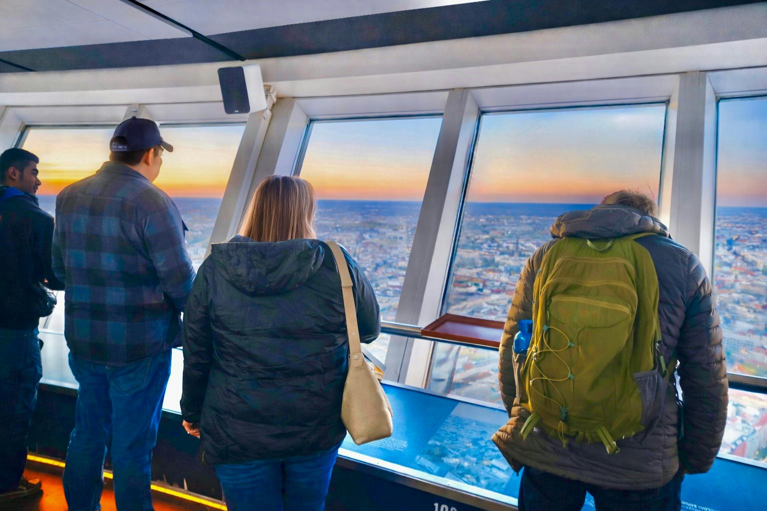 Folk står i nærheden af store vinduer på et observationsdæk og kigger ud på et bybillede ved solnedgang.