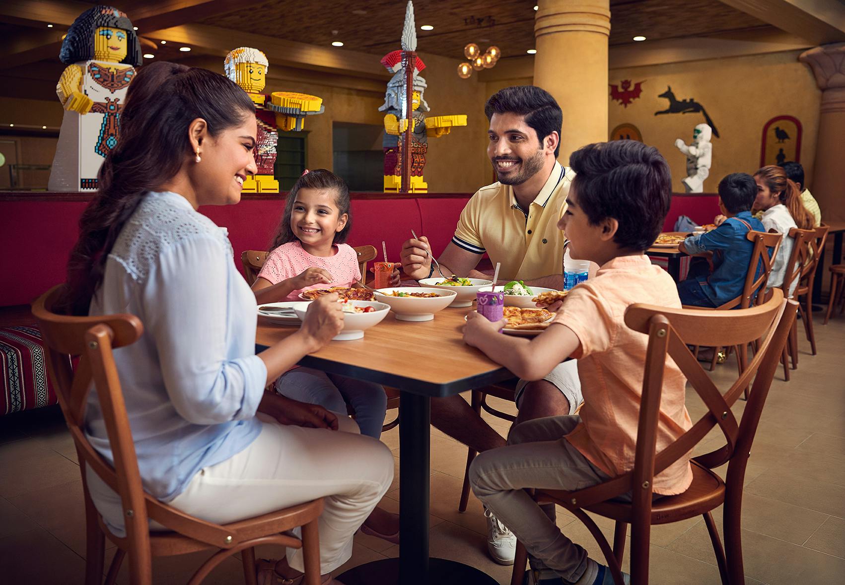 一家四口人微笑着在餐厅用餐，背景是乐高雕塑。