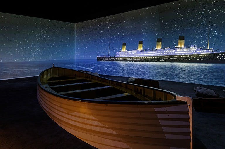 Un canot de sauvetage est placé à l'intérieur avec, en arrière-plan, une grande image illuminée du Titanic sur l'océan.