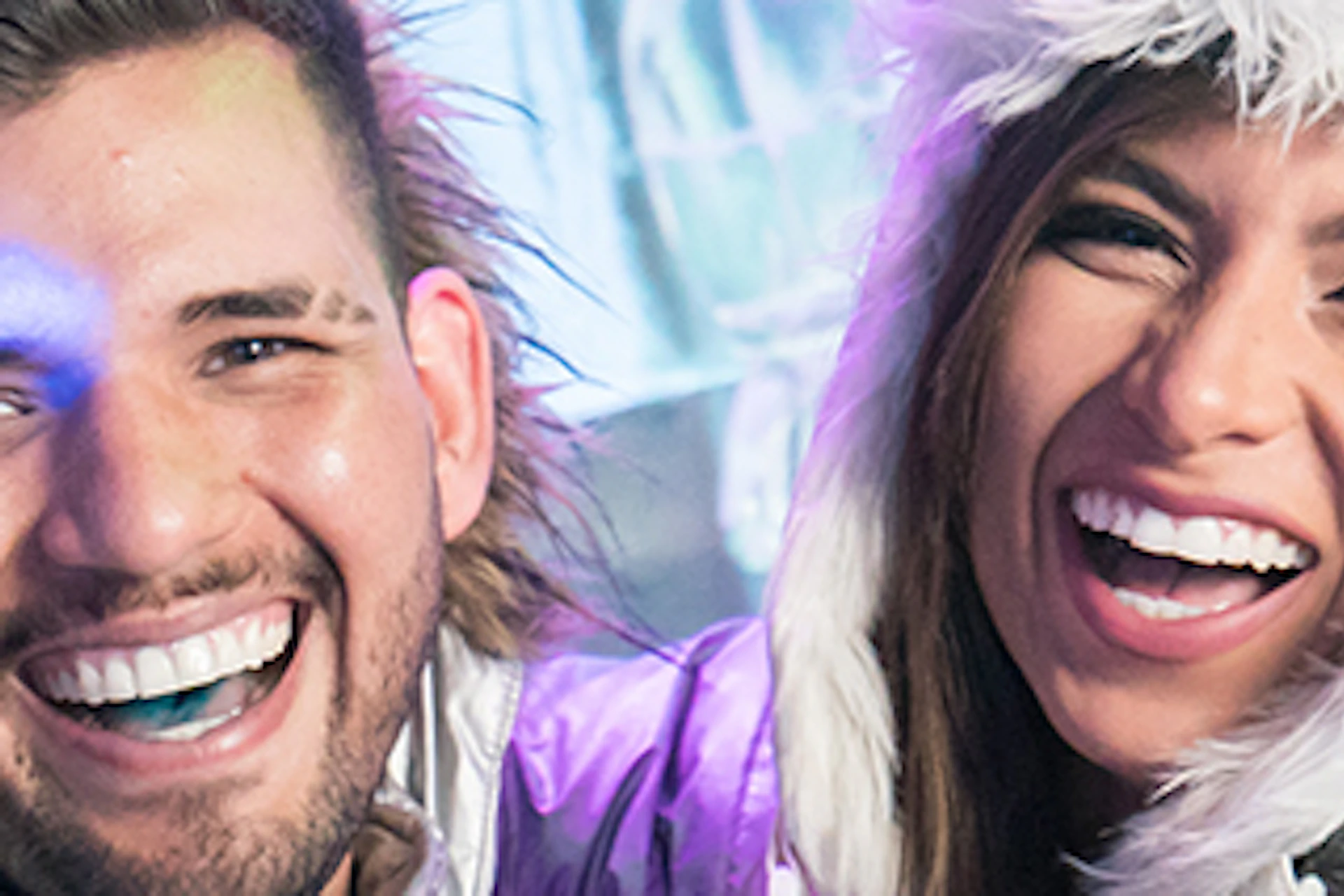 Two people in silver winter coats and furry hats smile and embrace in a setting with ice sculptures in the background.