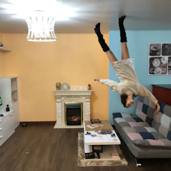 En kvinna verkar flyta upp och ner i ett vardagsrum med pastellfärgade väggar, en schackbrädessoffa och en TV som spelar naturscener.