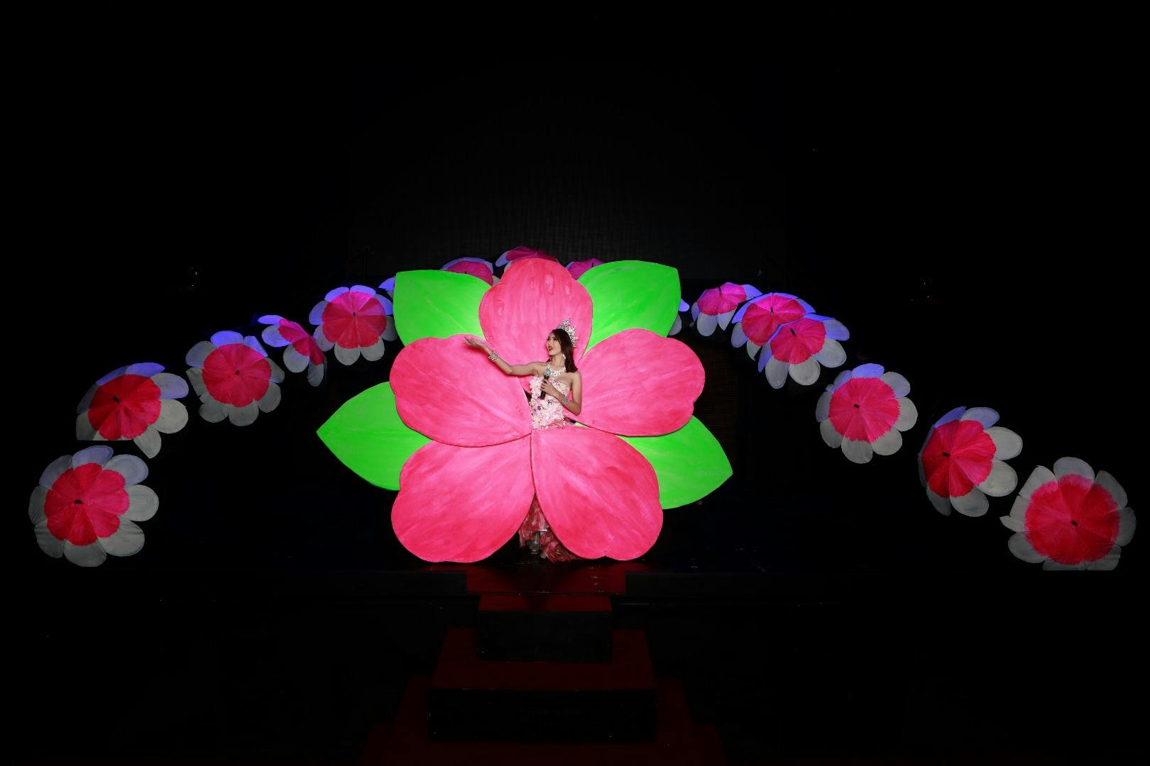 Una persona s'aixeca a l'escenari amb un disfressa que s'assembla a una gran flor rosa i verda, envoltada de decoracions florals més petites.