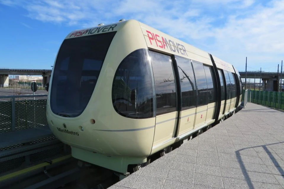 A beige and black "Pisa Mover" tram is stationed on tracks under a clear blue sky.