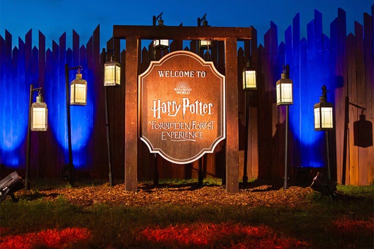 Ξύλινη πινακίδα που γράφει "Καλώς ήλθατε στο Harry Potter: Απαγορευμένη εμπειρία δάσους" με φανάρια, που περιβάλλεται από σκοτεινό φράχτη και μπλε φωτισμό.