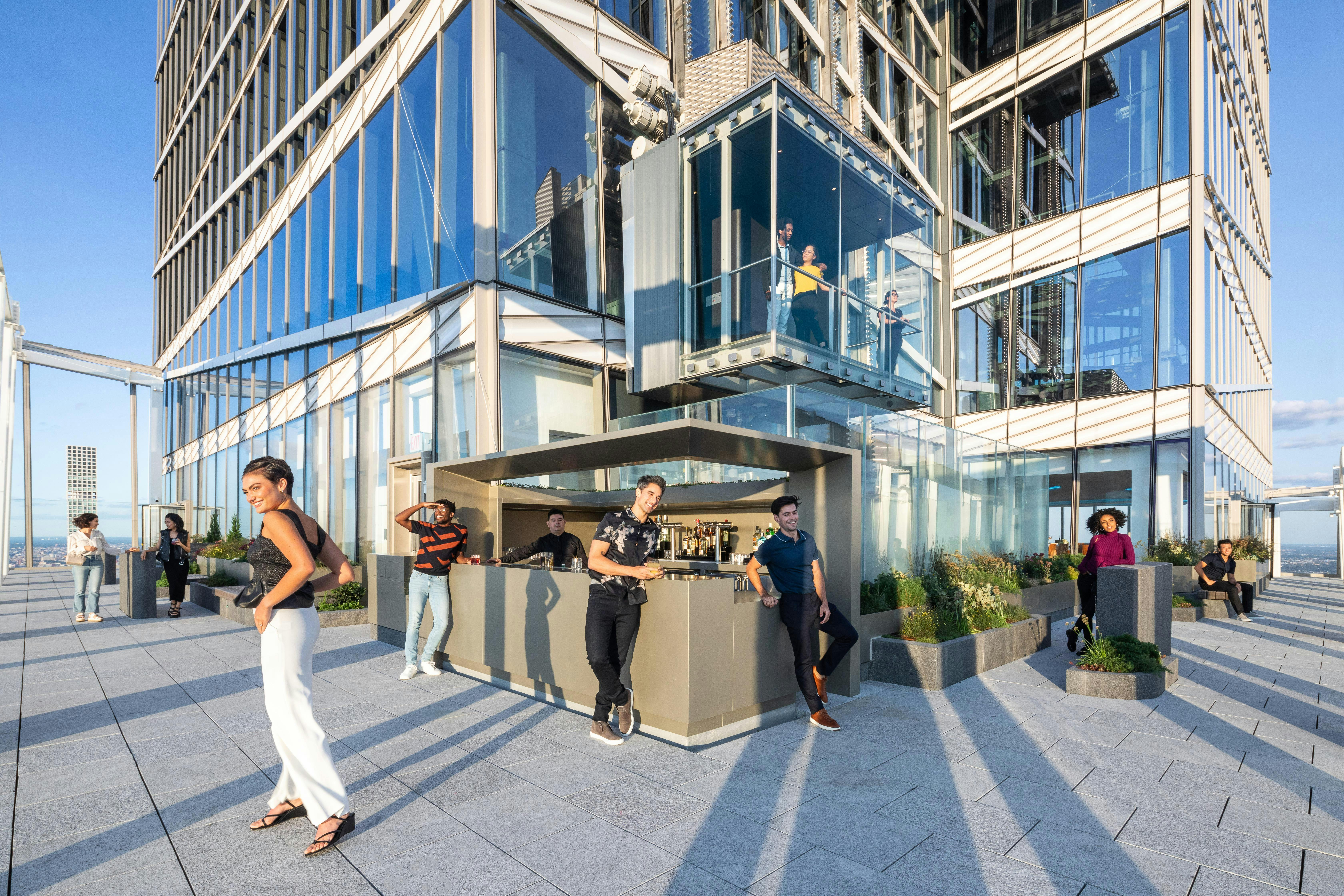 People relax and socialize on a modern rooftop terrace with seating and a bar, against a backdrop of gleaming glass buildings.