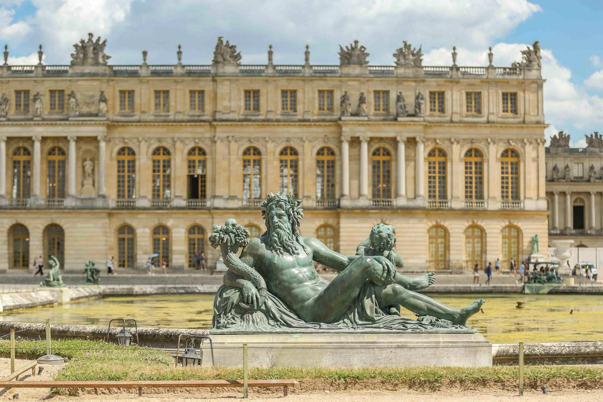 A large statue of a reclining man and a child in front of the Palace of Versailles with people walking in the background.