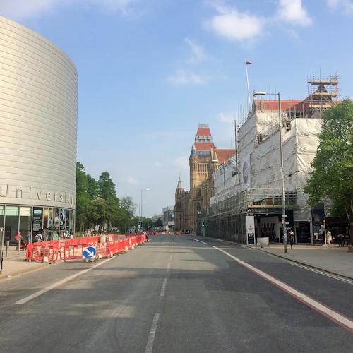 Gatuvy med en modern cylindrisk byggnad, en byggnad under byggnadsställningar, ett avlägset historiskt torn och en klarblå himmel.
