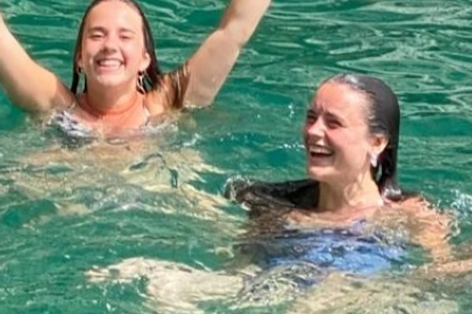 Three smiling people swimming in clear turquoise water near a rocky shoreline, raising their hands in a gesture of joy.