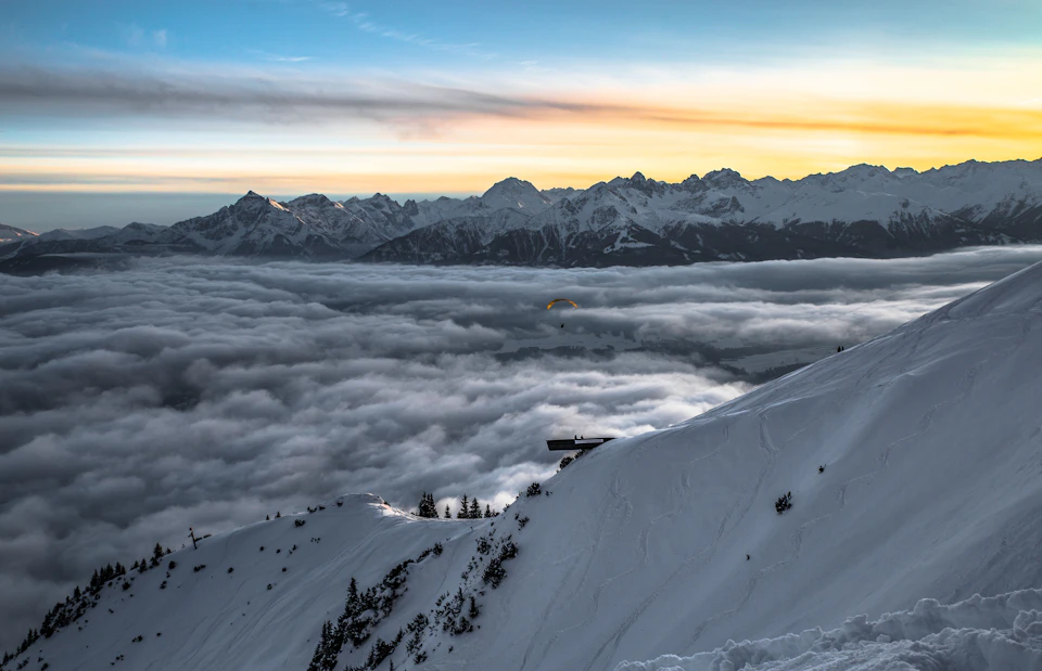 Snow-covered mountain slope with a wooden bench overlooking a sea of clouds below and distant mountains at sunrise.