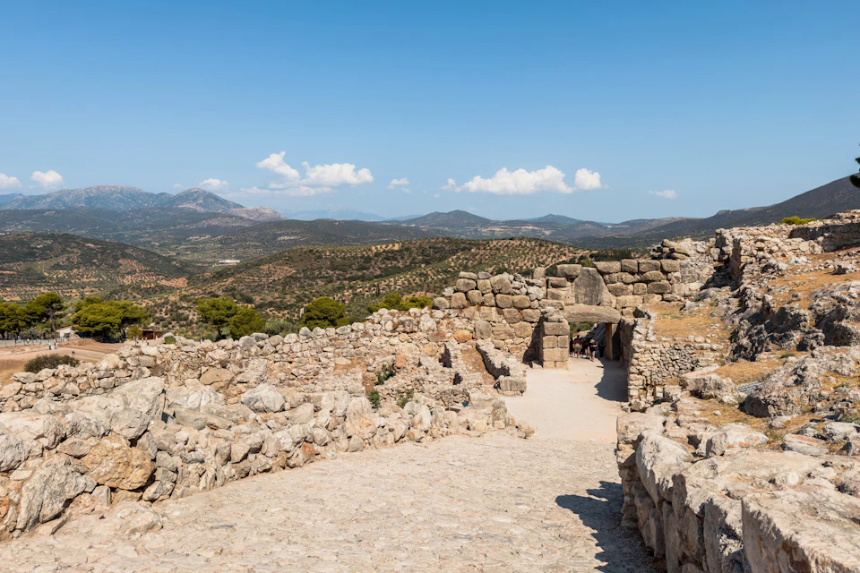 Ancient stone ruins with an arched gateway, surrounded by a hilly landscape under a bright blue sky.