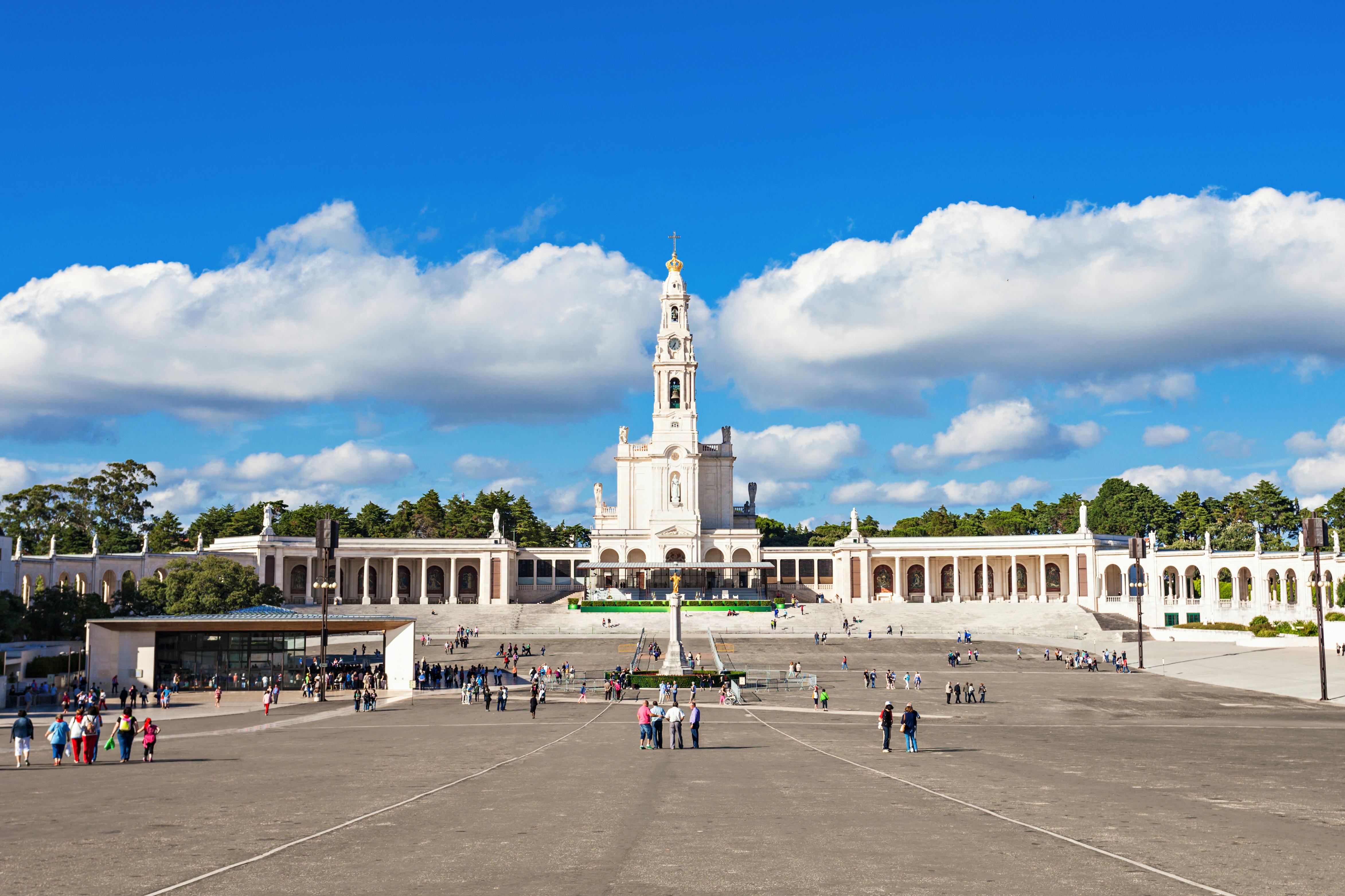 A large open plaza with scattered visitors, centered on a tall basilica with an ornate tower under a bright blue sky with clouds.