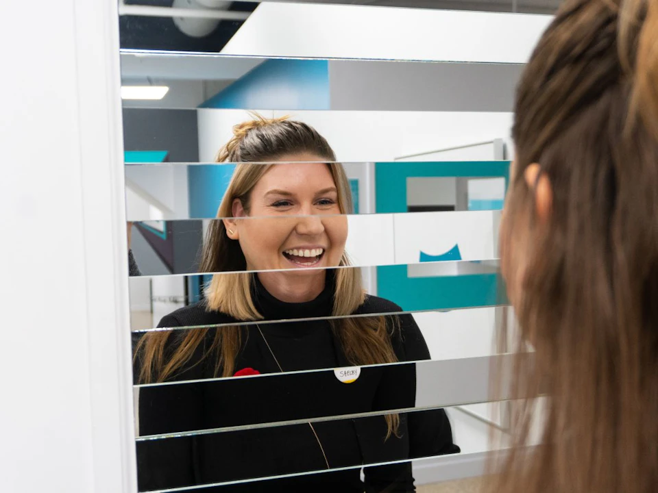 A smiling woman with a "Sarah" name badge behind a partially covered glass divider, interacting with another person.
