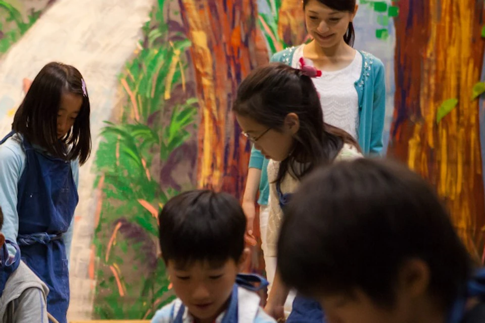 Children wearing aprons working on wooden blocks in a colorful indoor setting with painted scenery in the background.