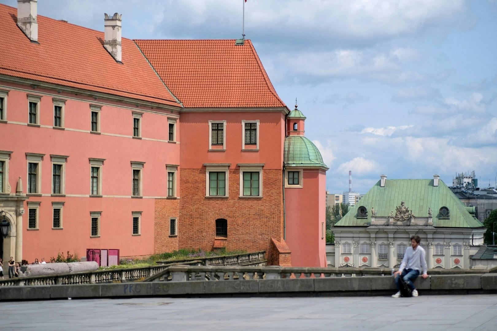 Ein historisches Gebäude mit orangefarbenen Wänden und einem grünen Kuppelturm, daneben ein weiteres Gebäude mit grünem Dach, unter einem blauen Himmel.