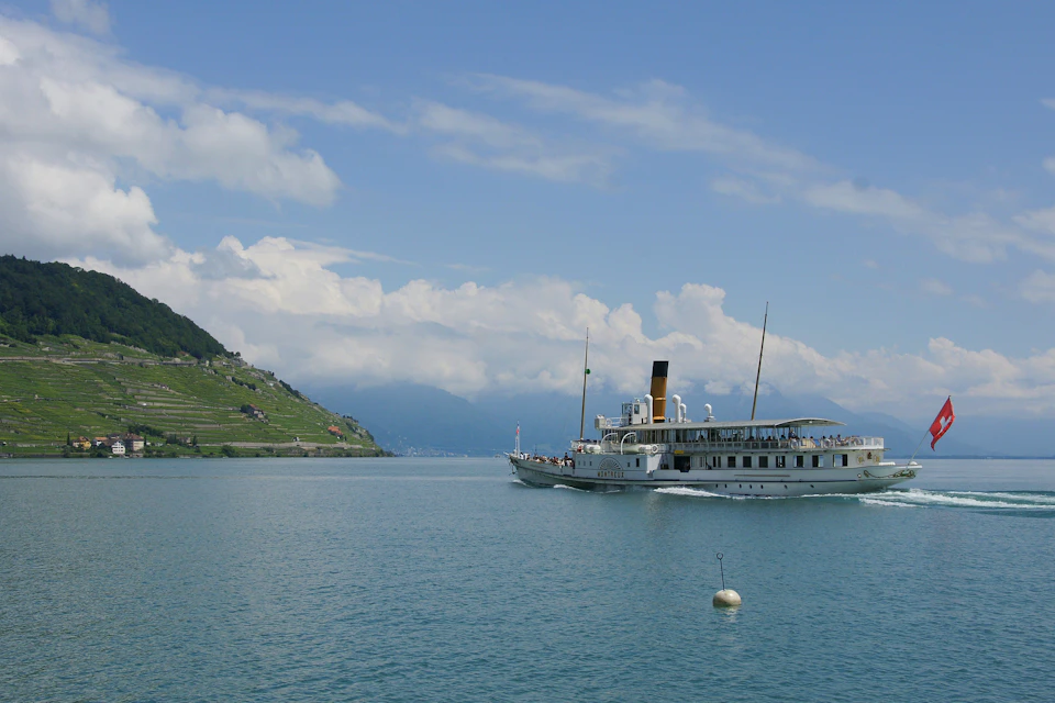 A steamboat sailing on calm water with a distant hillside covered in terraced vineyards and a partly cloudy sky.