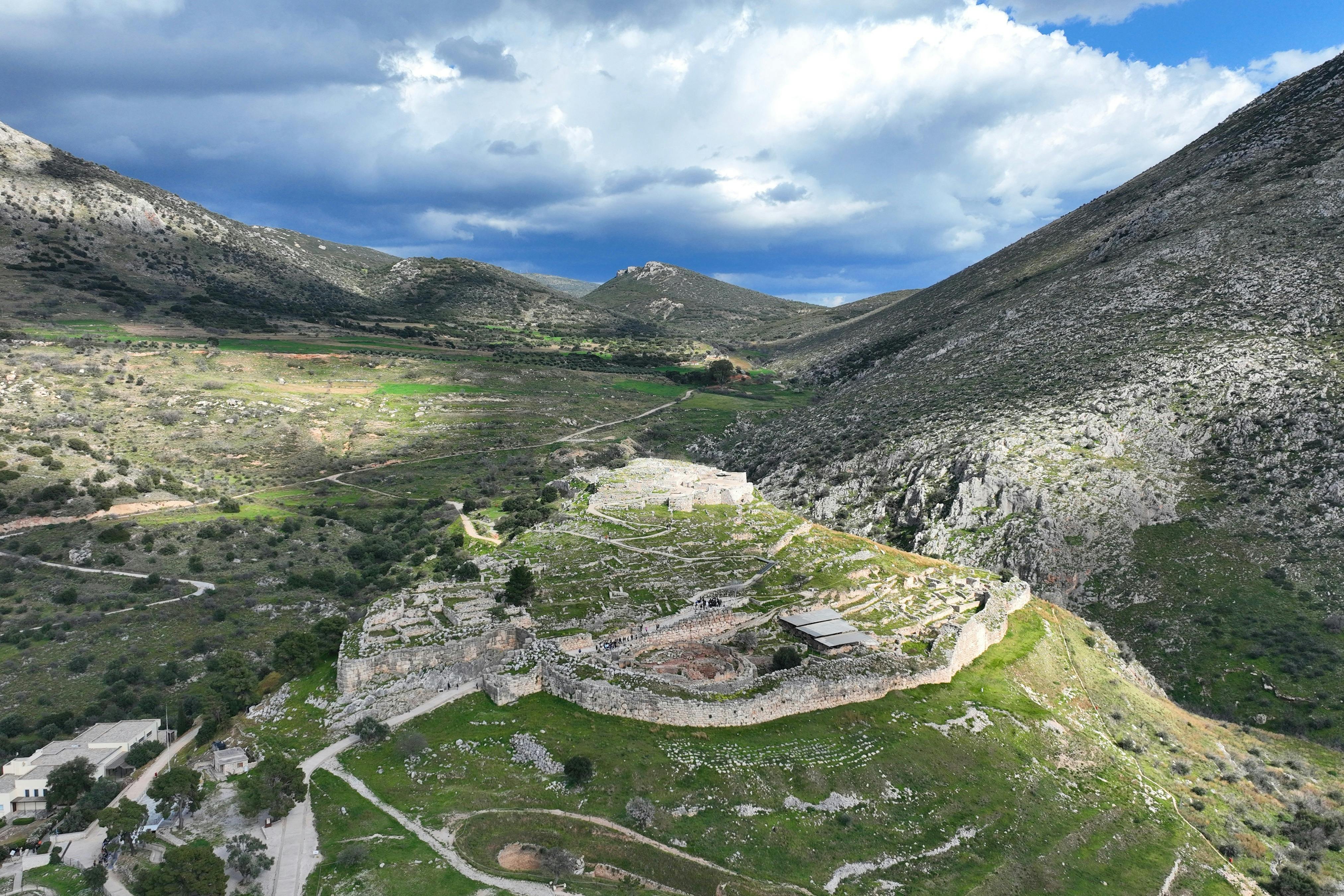 부분적으로 흐린 하늘 아래 푸른 언덕으로 둘러싸인 고대 석조 요새의 공중 전망.