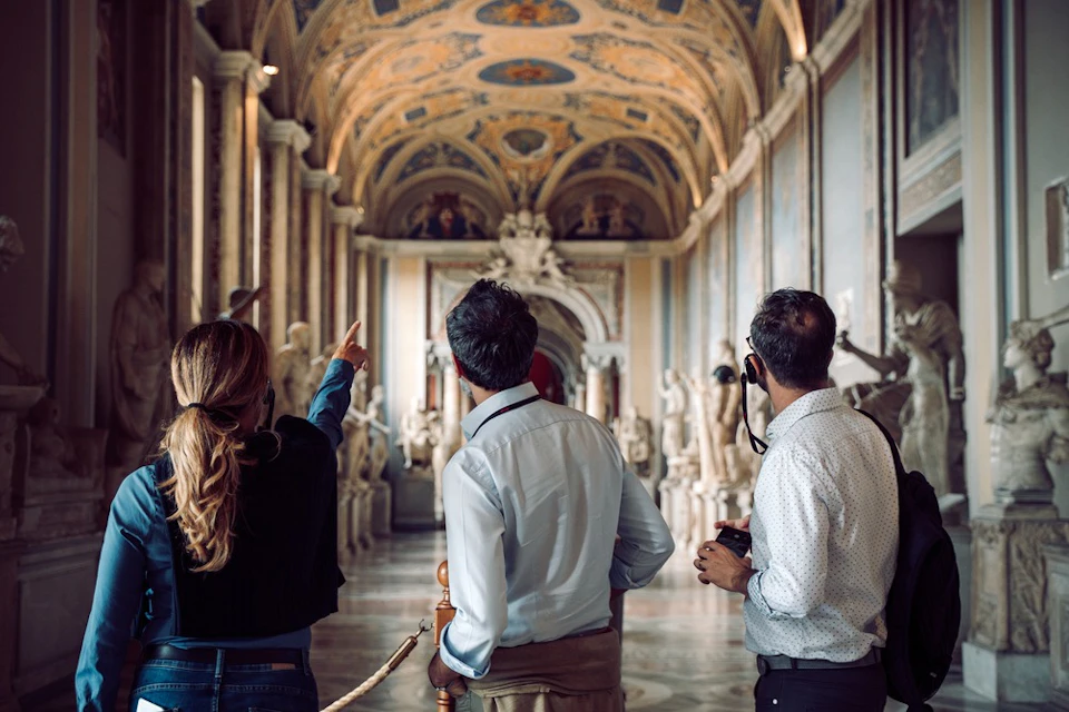 Three people in a museum hallway with ornate ceilings and classical sculptures, one pointing towards the ceiling.