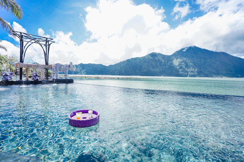 A pool with a floating tray holding drinks, a decorative elephant statue, and mountains in the background under a blue sky.