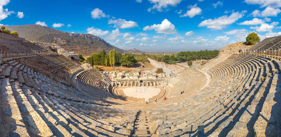 Ancient amphitheater with tiered stone seating, partially filled with people, surrounded by trees and mountains under a blue sky.