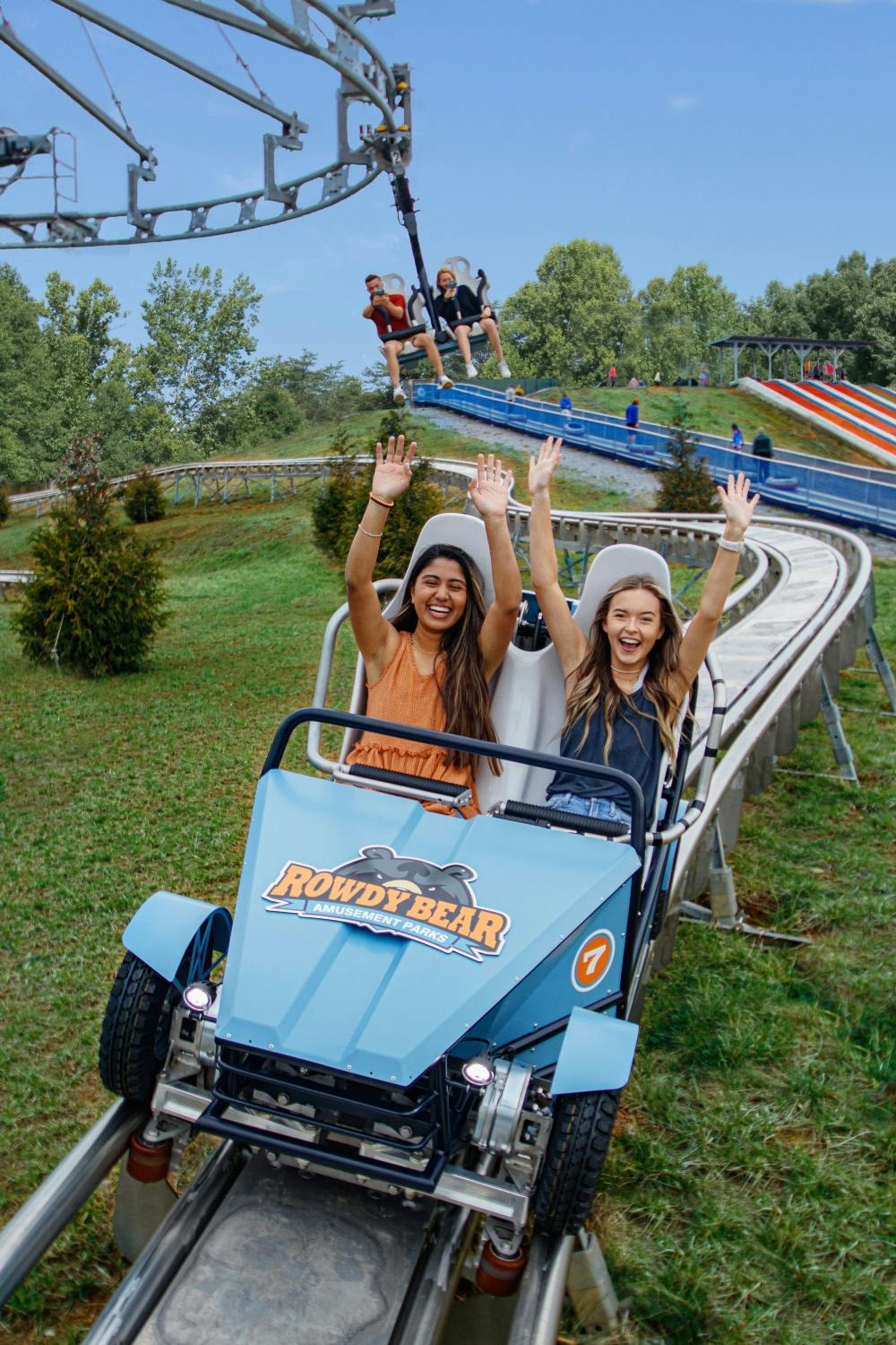 Two smiling women with raised arms ride a blue cart on a track at Rowdy Bear Amusement Park, with other riders in the background.