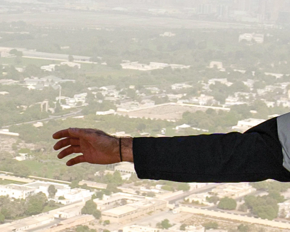 Two people in harnesses, arms outstretched, suspended high above a cityscape with skyscrapers and roads visible below.
