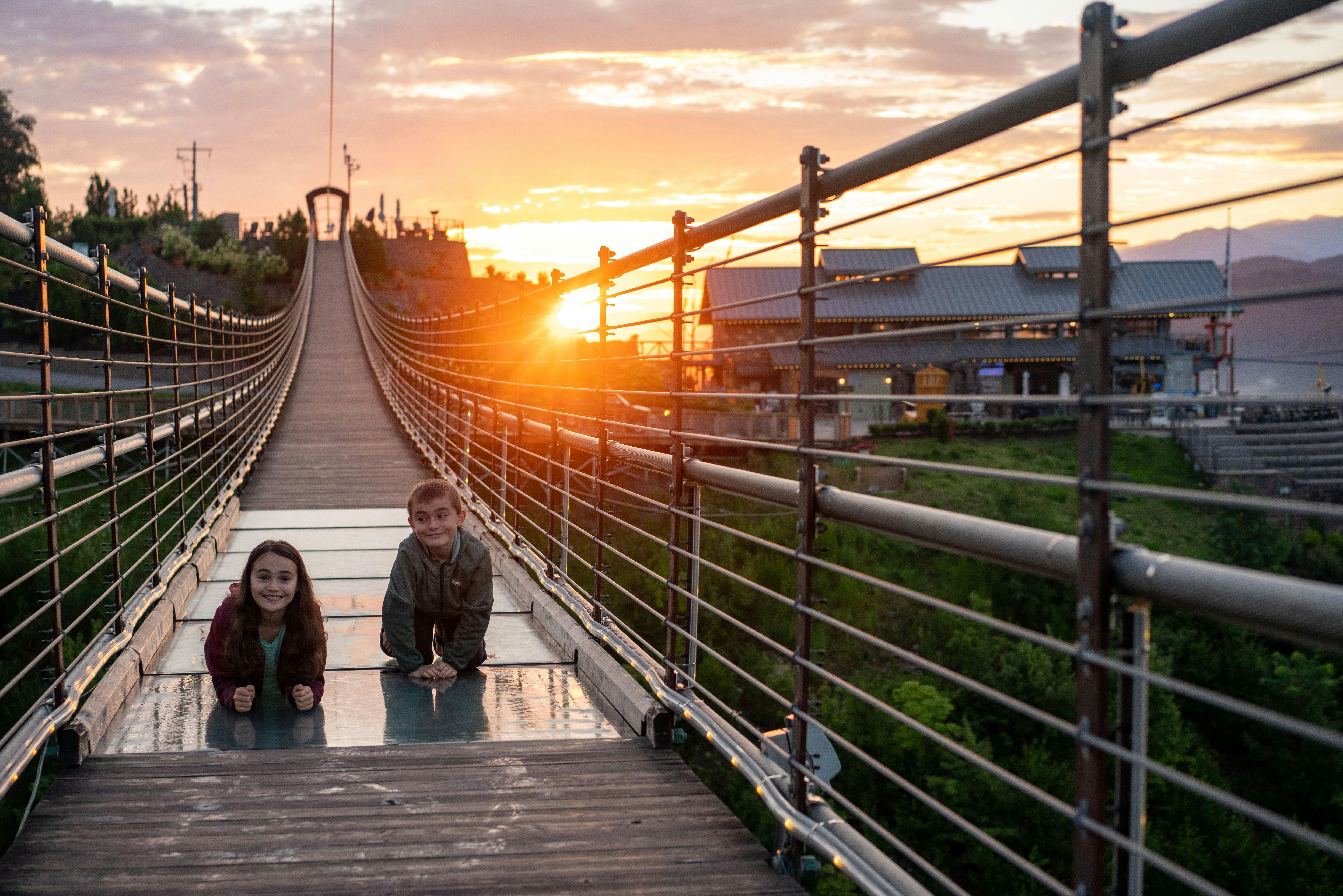 Dvě děti ležící na visutém mostě při západu slunce, usmívající se do kamery s budovami a stromy v pozadí.