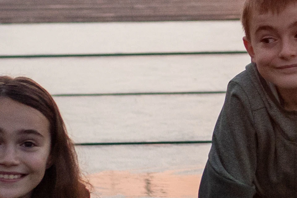 Two children lying on a suspension bridge at sunset, smiling at the camera with buildings and trees in the background.