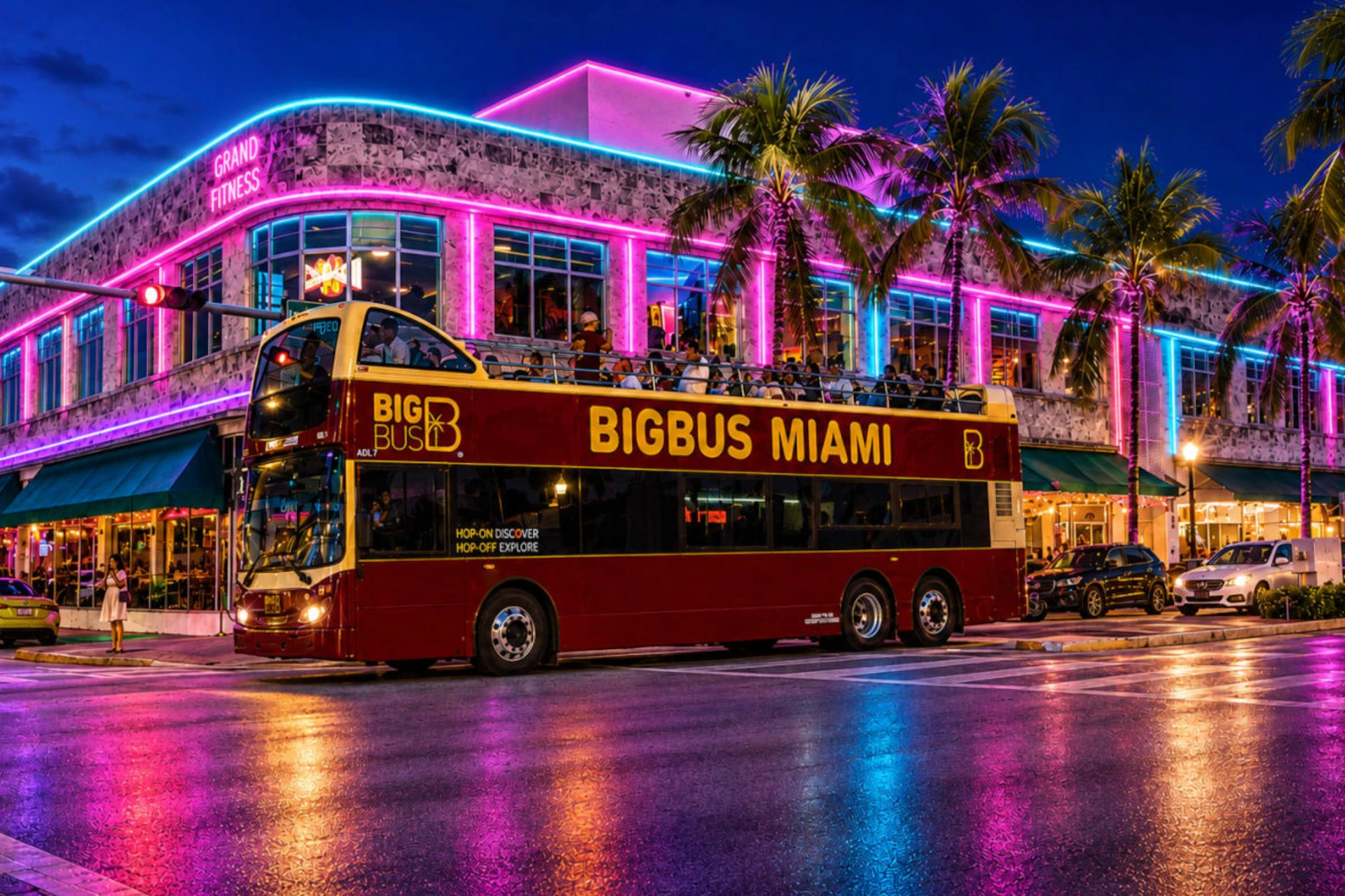 A red double-decker bus labeled 'Big Bus Miami' parked on a street at night, with a neon-lit building and palm trees in the background.