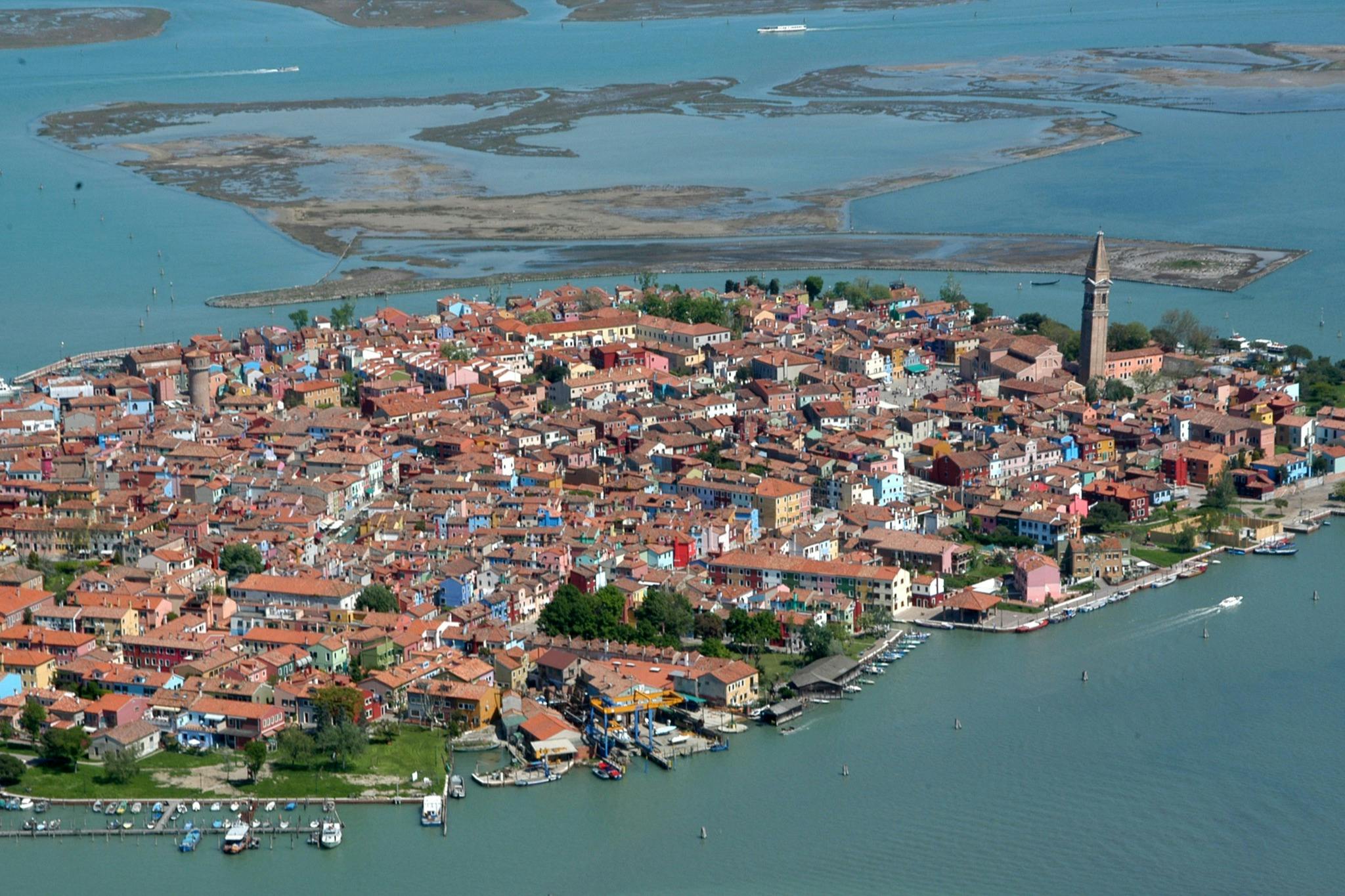 水に囲まれたカラフルな町の空からの眺めで、傾斜した鐘楼と海岸線にある小さなボートを特徴としています。