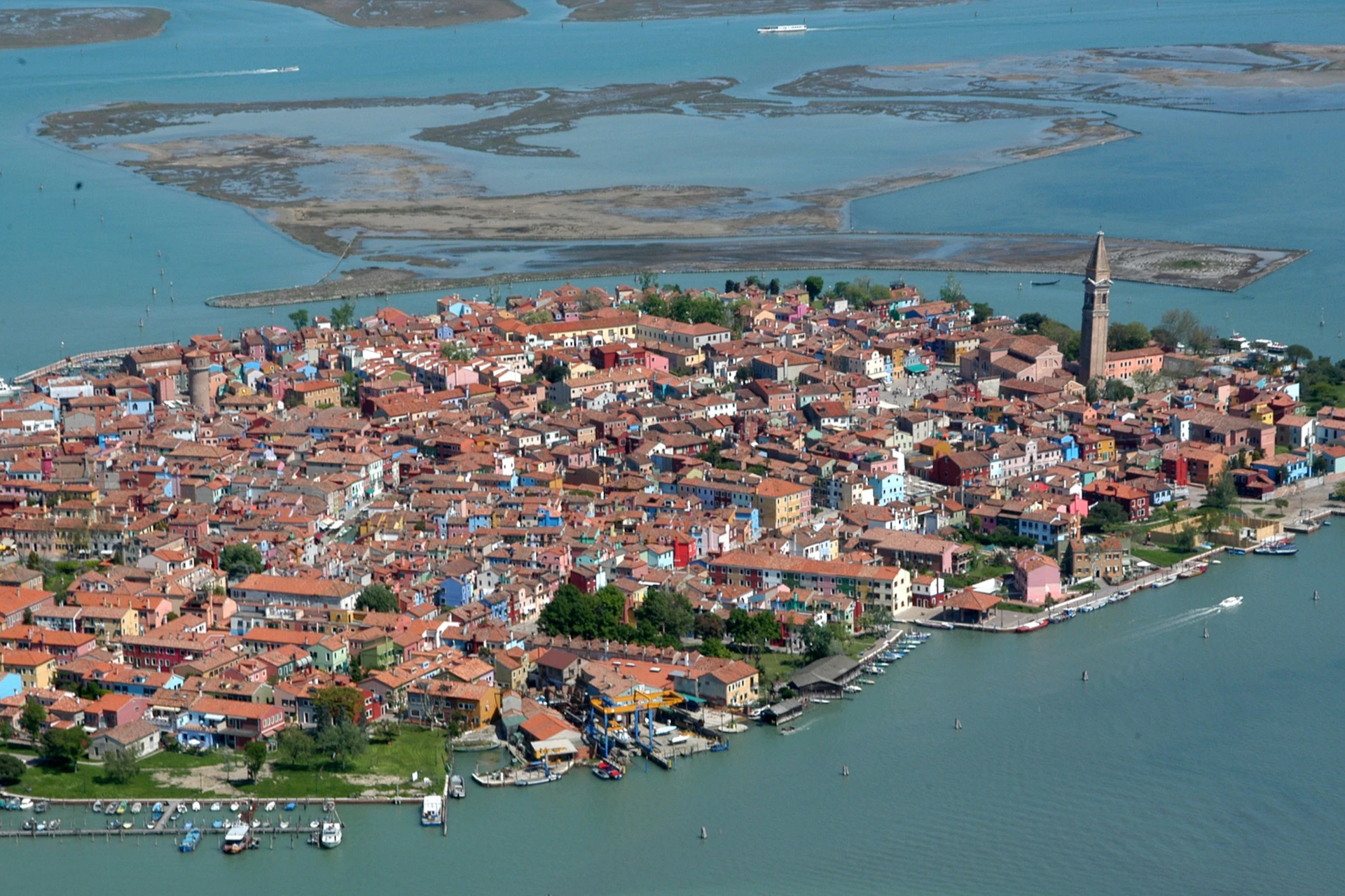 An aerial view of a colorful town surrounded by water, featuring a leaning bell tower and small boats at the shoreline.