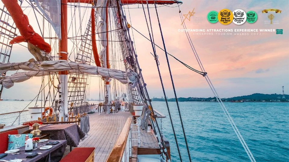 A sailboat on calm waters at sunset, featuring wooden deck, masts, rigging, and distant shoreline. Text: "Outstanding Attraction, Singapore Tourism."