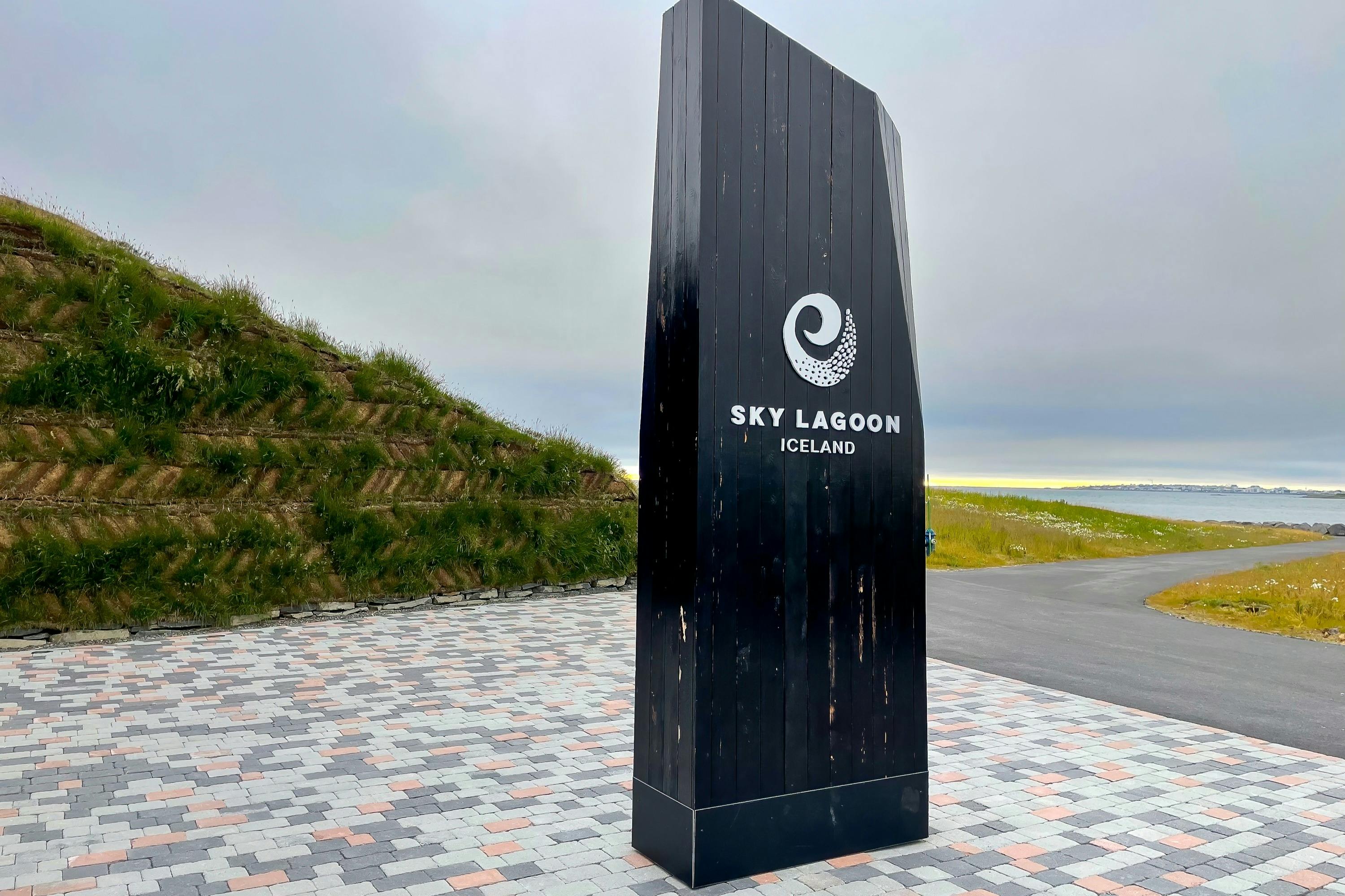 Černá cedule s logem „Sky Lagoon Iceland“, stojící na vzorovaném chodníku. Trávou pokrytý kopec a v pozadí zatažená obloha.
