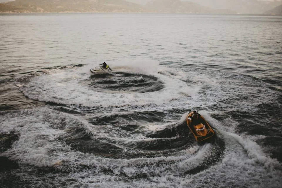Two people riding jet skis, creating circular waves on a calm lake with mountains in the background.