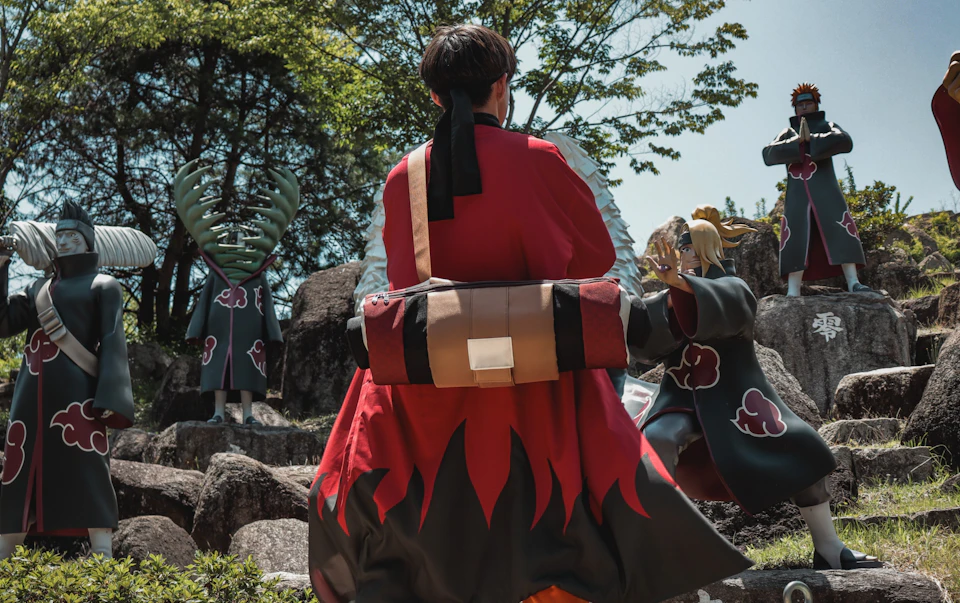 A person in a red and black outfit with a rolled-up cloth bag on their back, standing outdoors near statues and trees.
