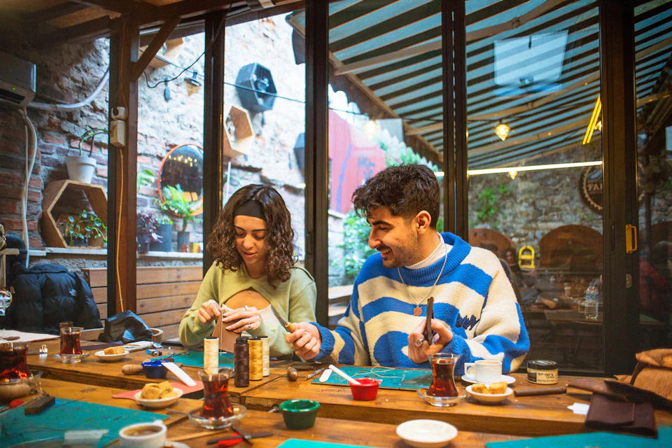 Two people sitting at a wooden table in a cozy workshop setting, engaged in a craft activity with tools and materials.