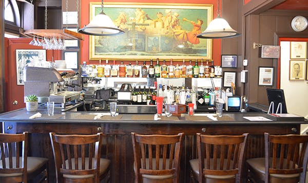 Un bar con una fila di sgabelli di legno vuoti, varie bottiglie di alcolici su scaffali, lampade appese e un murale sopra il bancone.