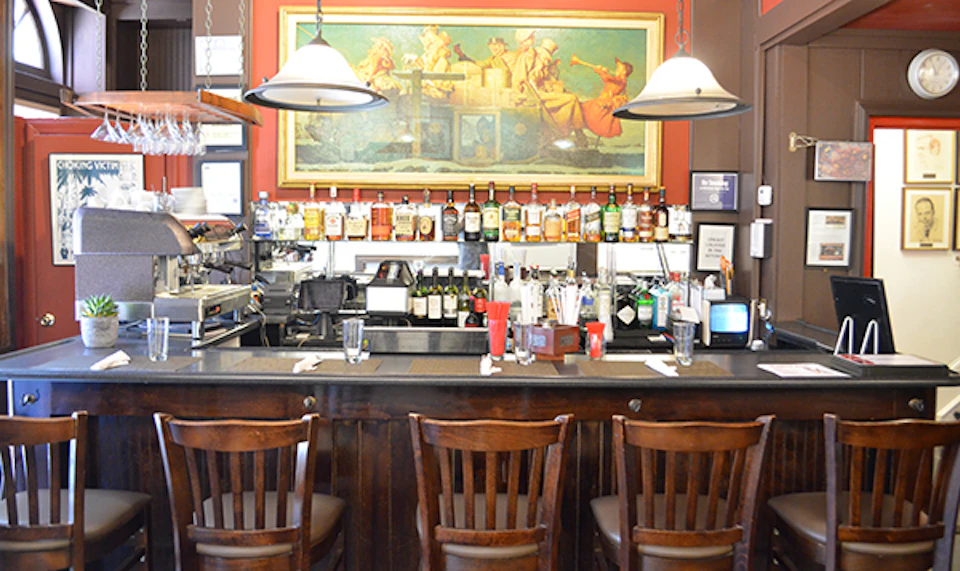 A bar with wooden stools, a variety of bottled drinks behind the counter, and a large Renaissance painting on the wall.