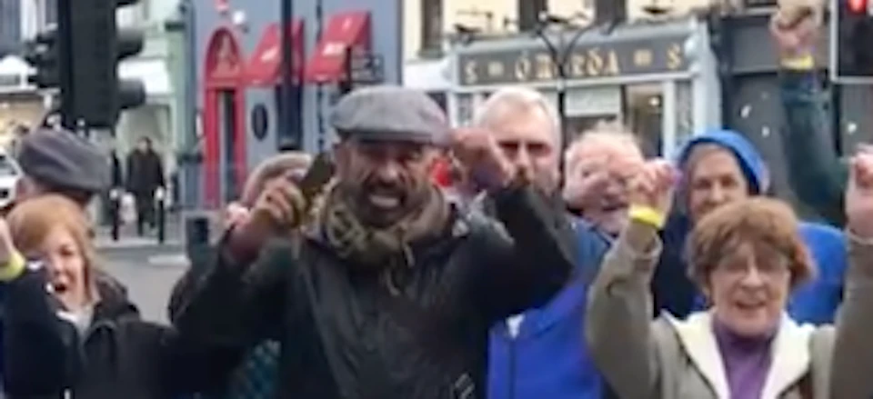 A group of people on a city street raise their hands excitedly while interacting with a man holding a paper bag.