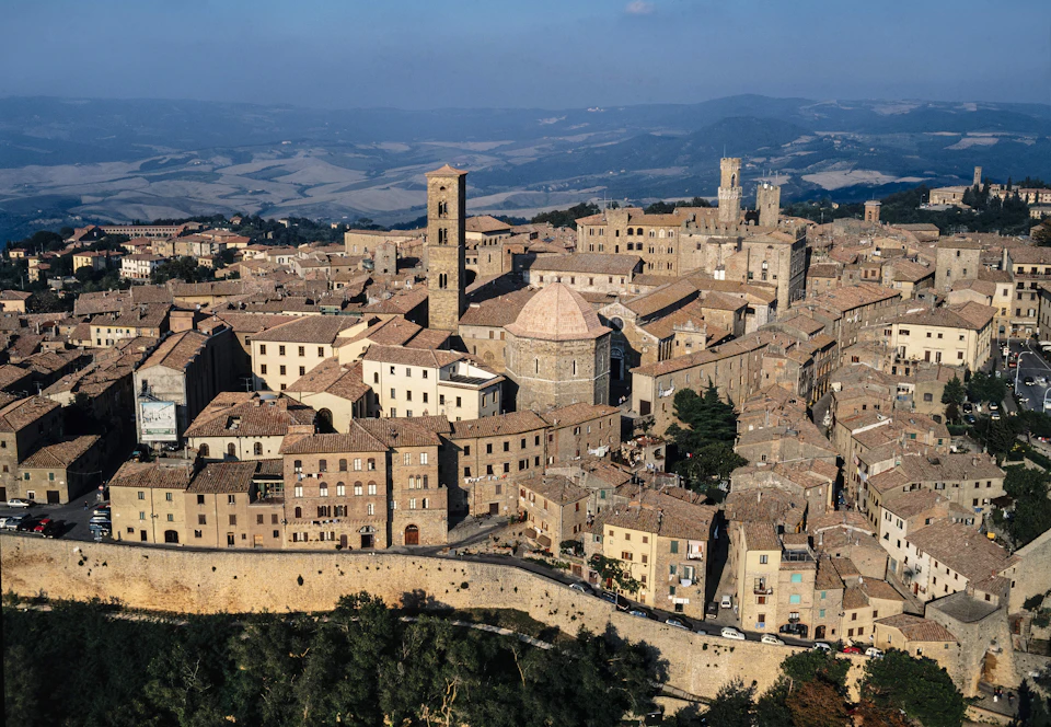 Aerial view of an old town with brown-roofed buildings, a tall bell tower, and surrounding hills in the background.