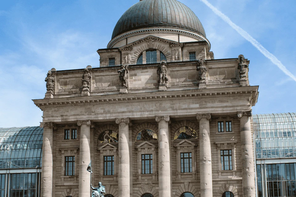 A historic building with columns, statues, and a domed roof under a blue sky with a contrail visible.