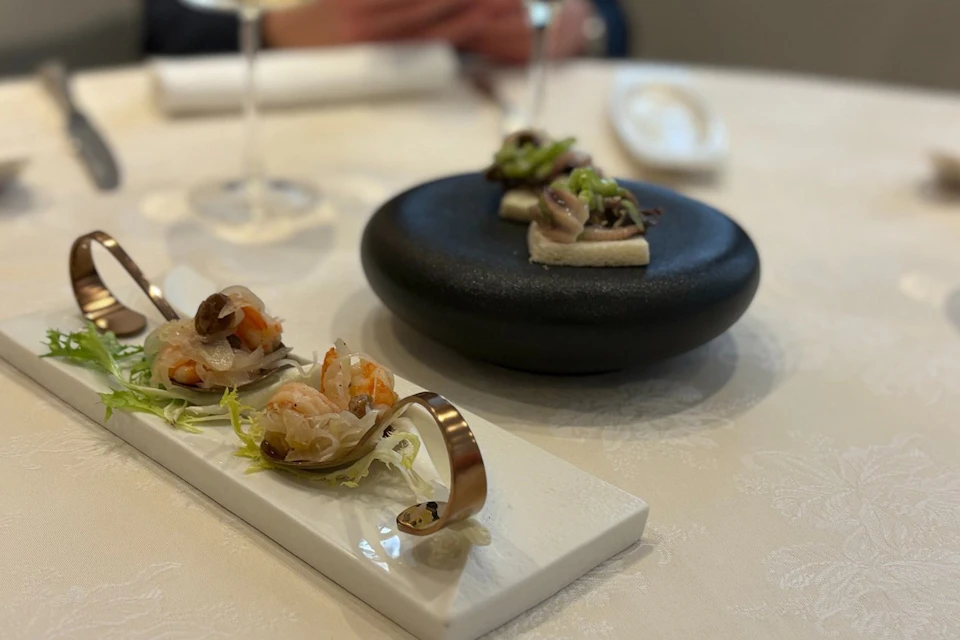 A refined dish featuring small gourmet servings on a white platter and a black stand, set on a white tablecloth.