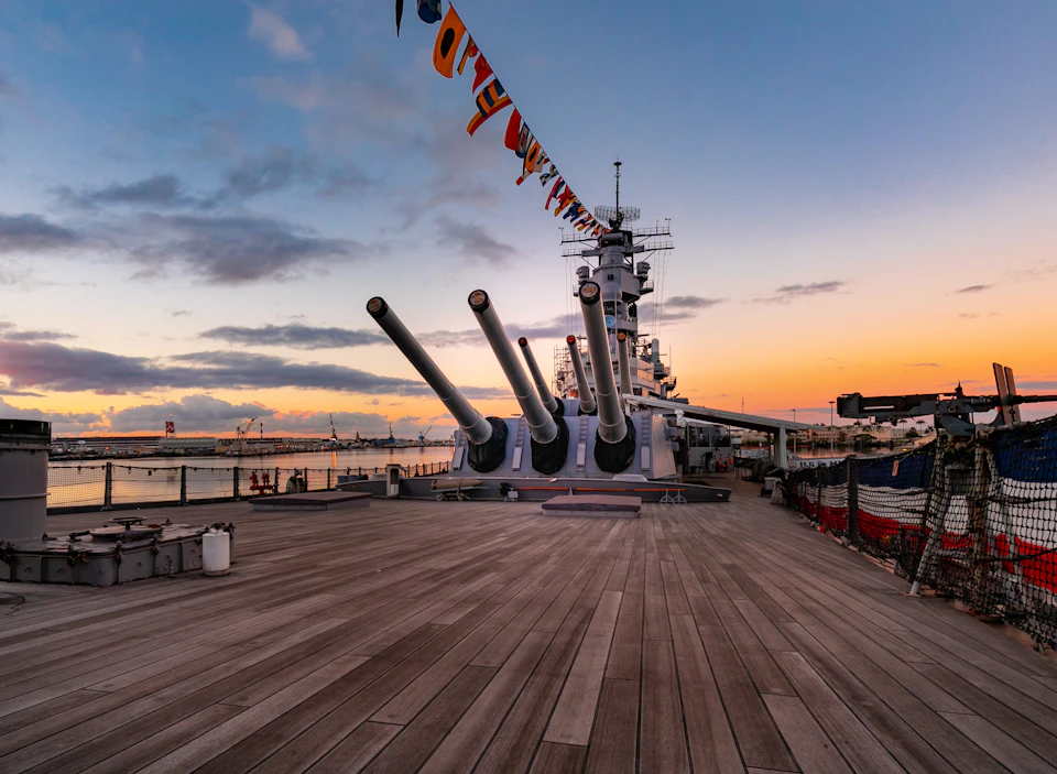 A battleship deck at sunset with three large cannons aimed forward, nautical signal flags hanging above.