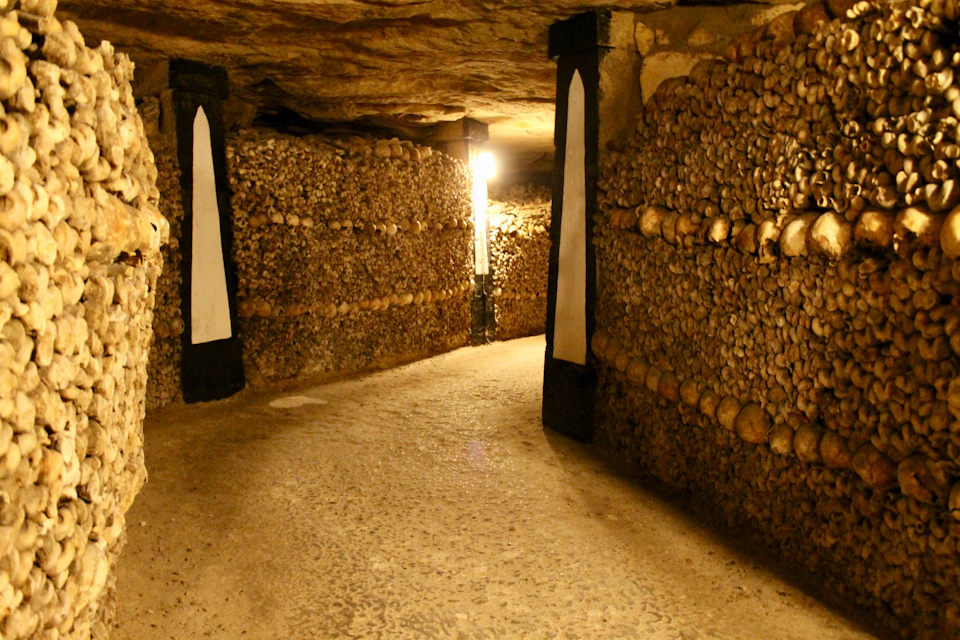 A dimly lit underground passage with walls made of neatly stacked bones and skulls, featuring decorative black pillars.