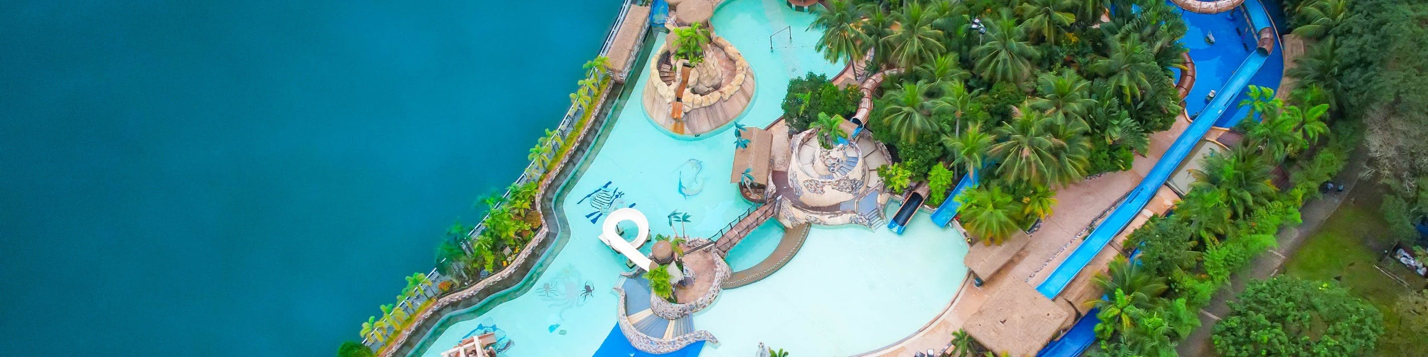 Vue aérienne d'un parc aquatique comprenant des bassins, des toboggans, des passerelles et des décorations sur le thème des tropiques, le tout entouré de verdure.