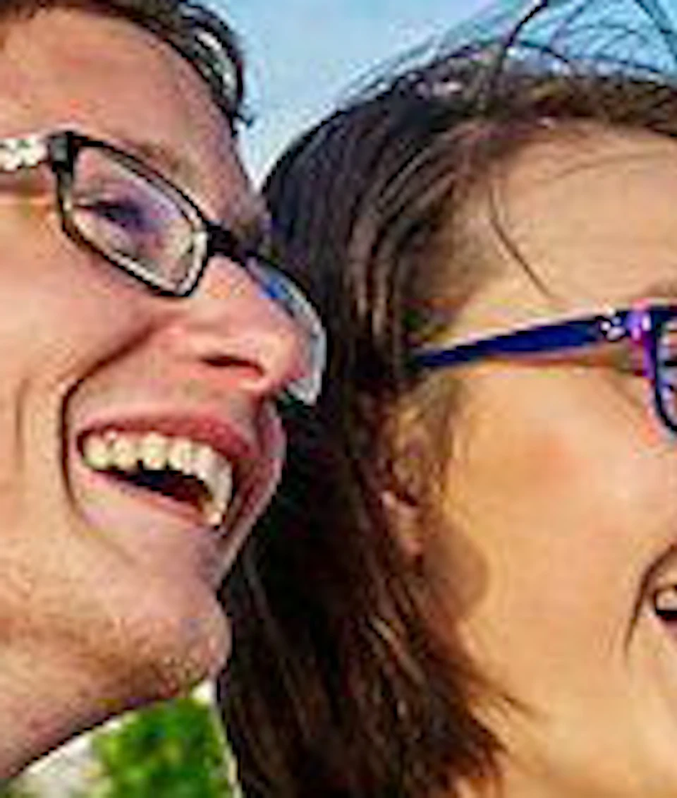 A smiling couple with glasses sitting on a boat, holding a glass of champagne, with a cityscape and river in the background.