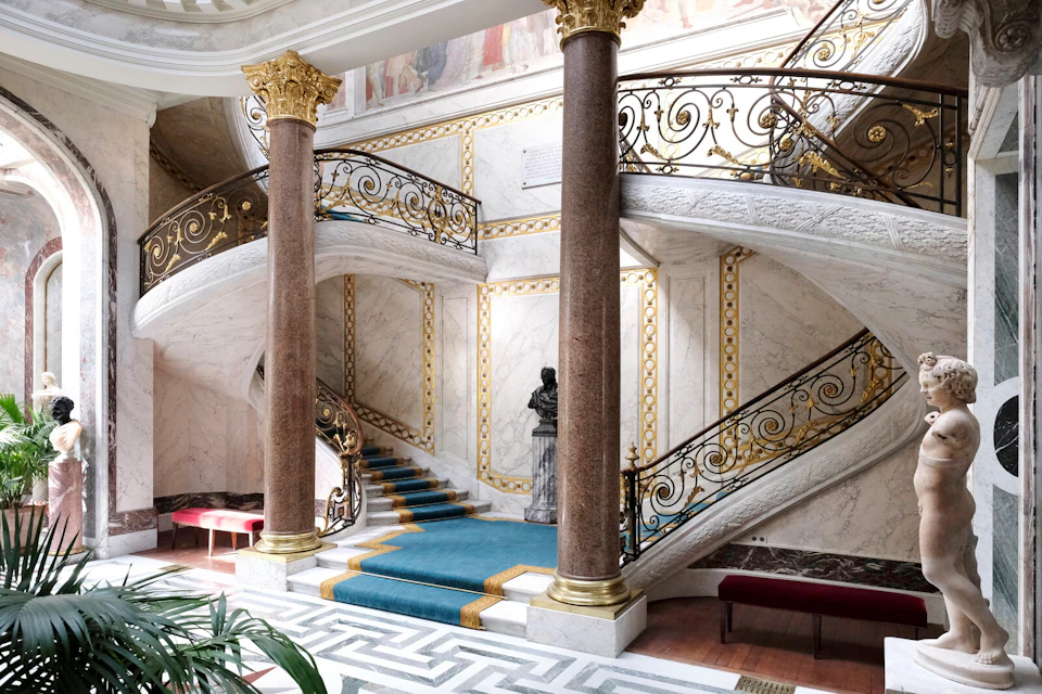A grand staircase with ornate railings, blue carpet, columns, and wall art in an elegant, classical interior setting.