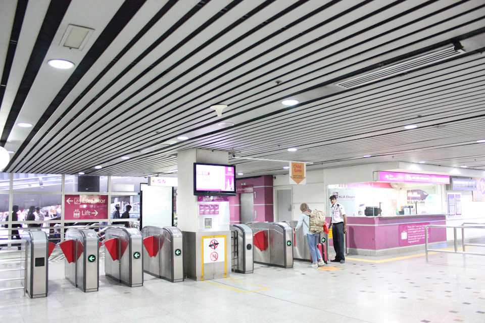A subway station entrance with turnstiles and a ticket booth. A person interacts with staff next to a sign for an elevator.