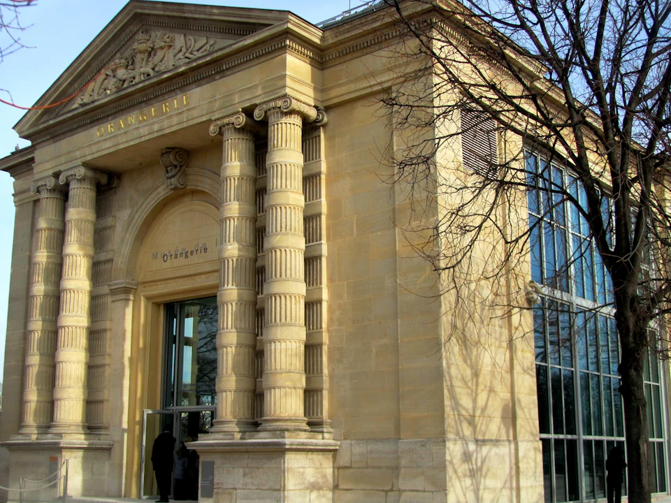 Ornate stone building entrance with tall, fluted columns, and "Orangerie" above the door, next to large glass windows and a leafless tree.