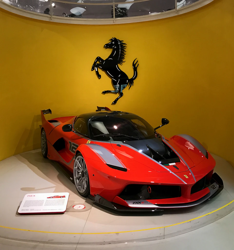 A red Ferrari sports car displayed against a yellow wall with a black prancing horse logo. Information plaque on the floor.