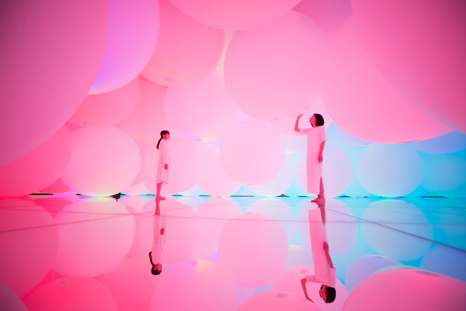 Two people in white dresses stand among large, glowing pink and purple balloons on a reflective floor.